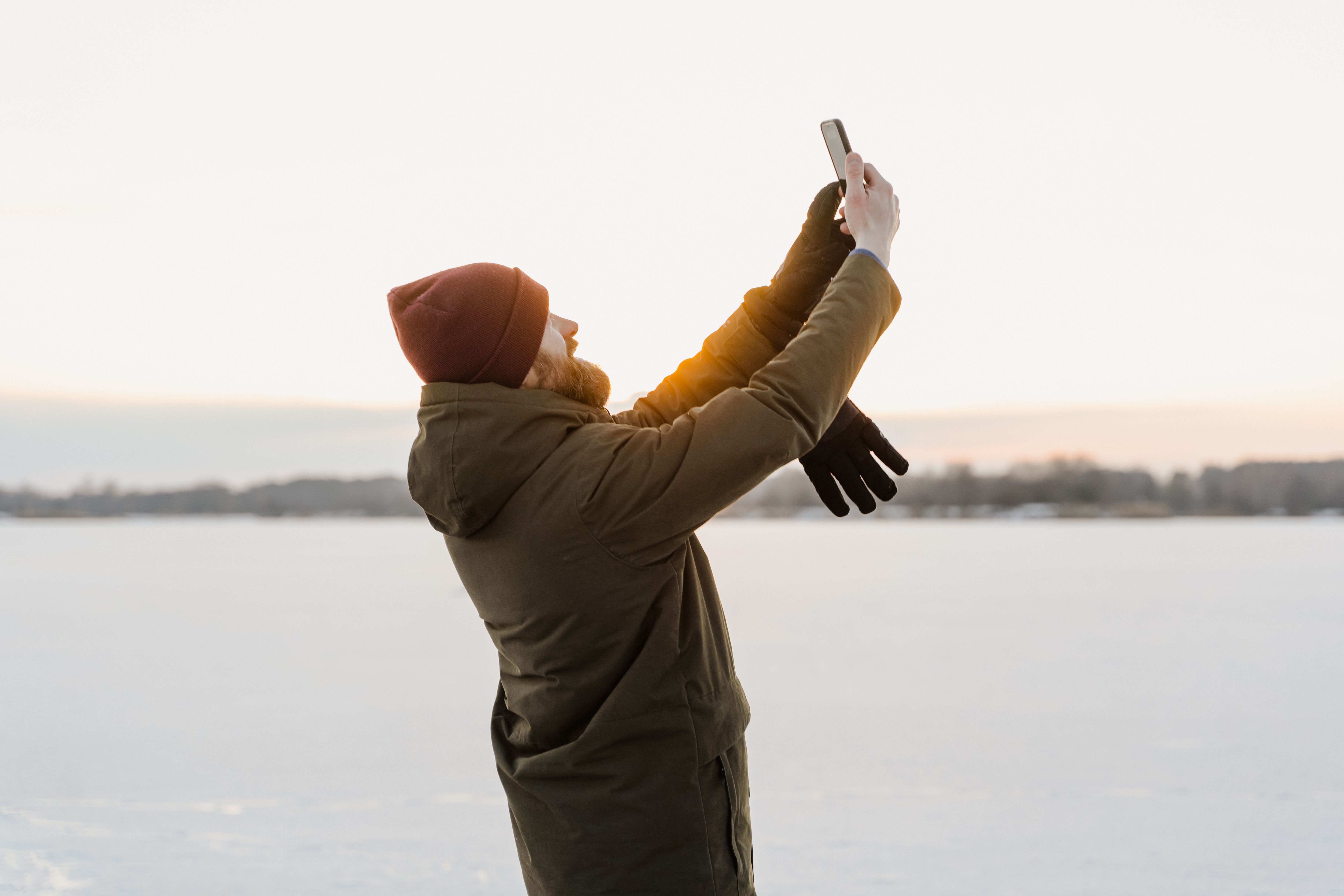 Bearded man taking a selfie against a snow-covered background during sunset.