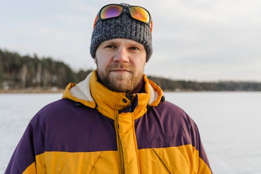 Portrait of a bearded man in warm winter clothing standing in a snowy landscape, wearing a beanie and sunglasses.