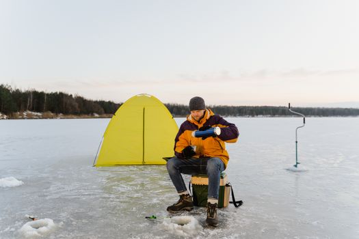 A man ice fishing next to a tent on a frozen lake during winter.
