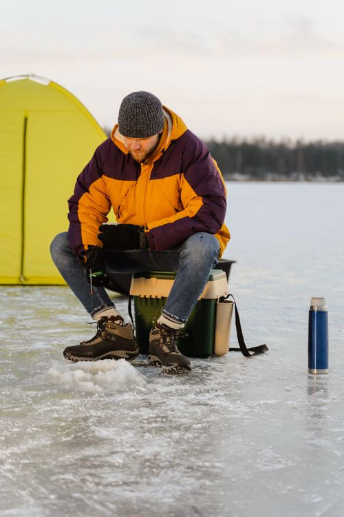 Free Man ice fishing on a frozen lake in winter gear near a tent and thermos. Stock Photo