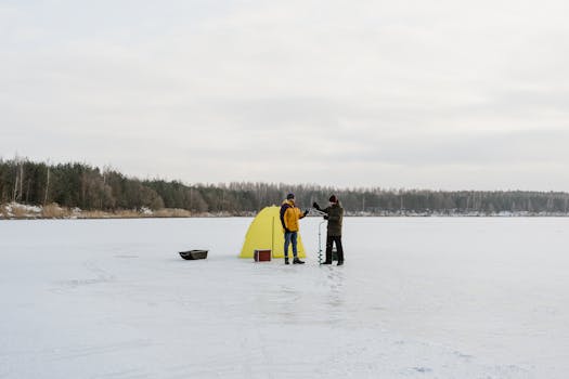 Two men ice fishing on a frozen lake with a yellow tent, surrounded by a snowy landscape.