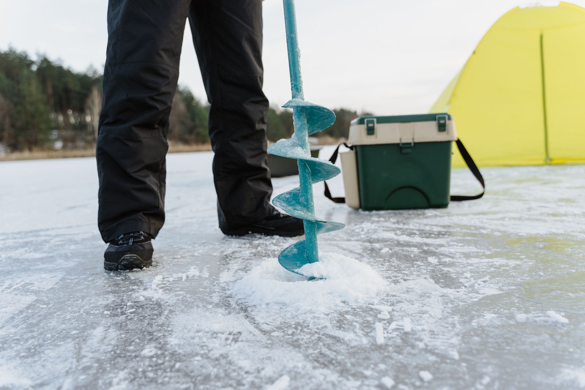 Person setting up ice drill for fishing on a frozen lake with equipment nearby.