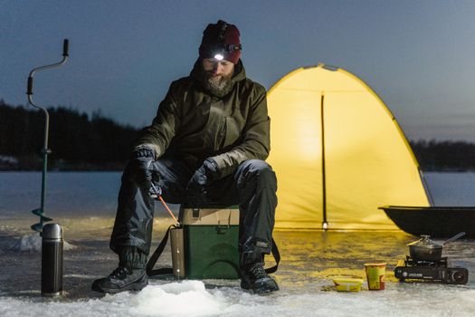 A man ice fishing by a camping tent on a frozen lake at night, embodying winter adventure.