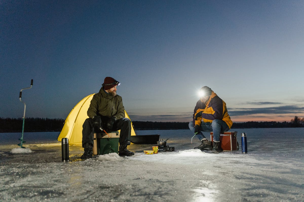 Two men ice fishing on a frozen northern lake at dusk, tent behind them — the classic pimpelfiske scene of Swedish Lapland winter