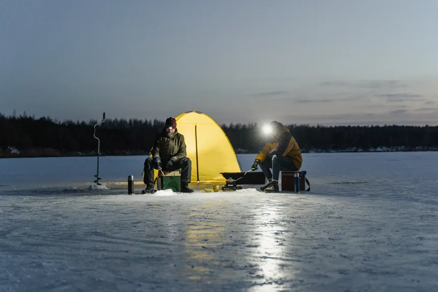 Two people ice fishing by a glowing tent on a frozen lake at night.