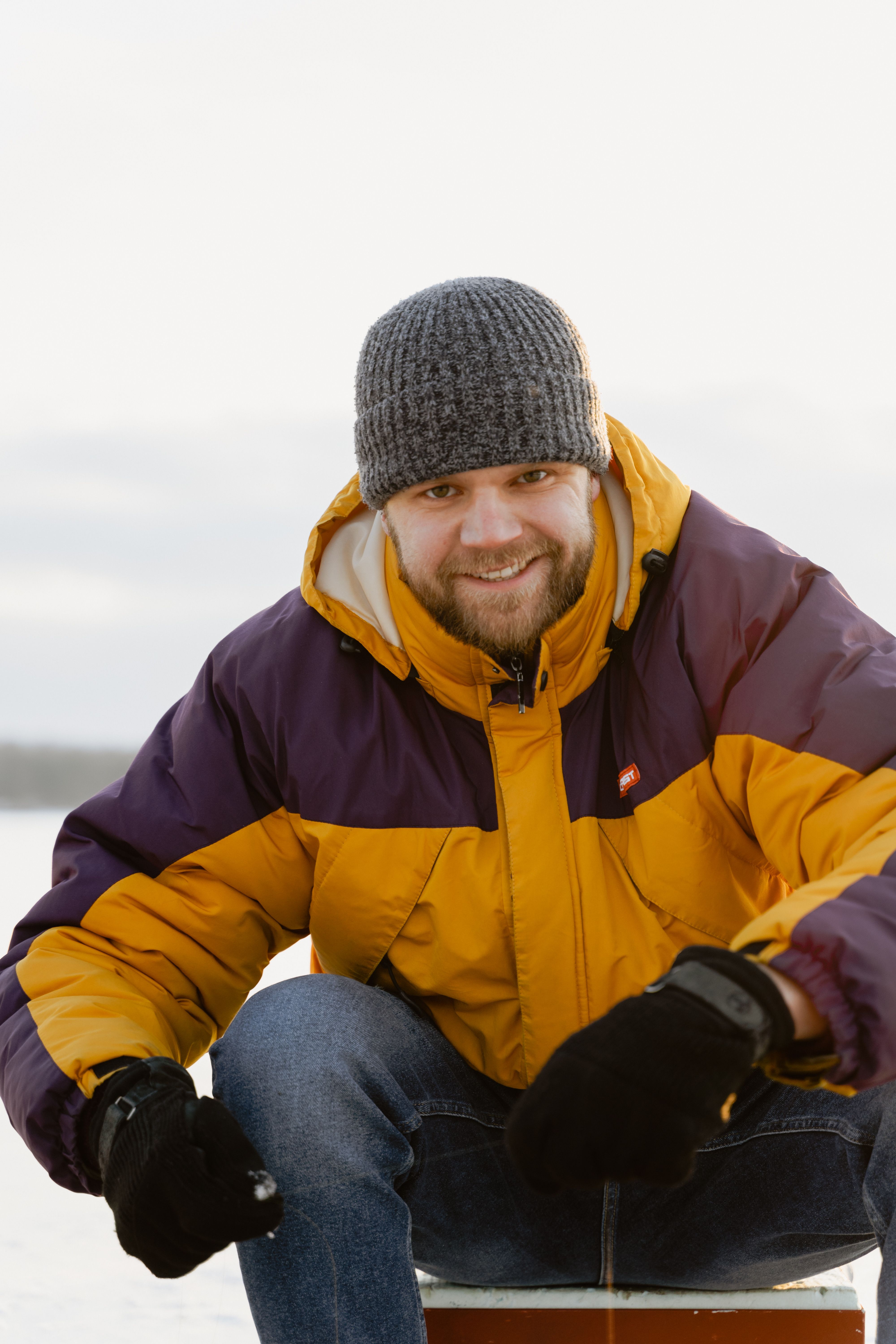 A cheerful man wearing winter clothes, sitting and smiling outdoors.