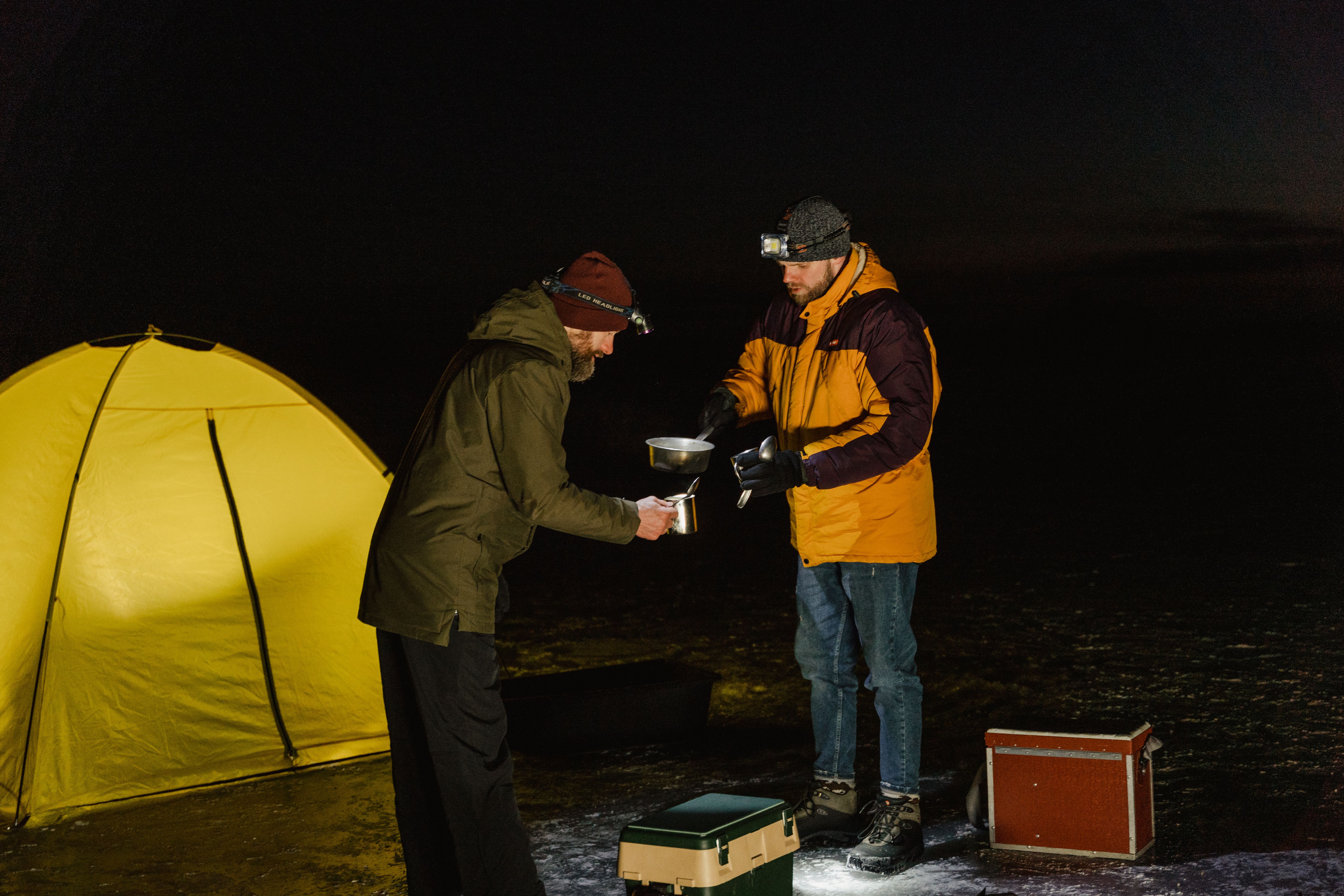 Two men camping at night in winter, sharing a meal by their tent.