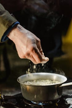 Close-up of a hand sprinkling herbs into a steaming saucepan during cooking.