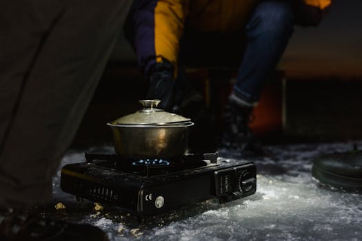 A portable stove with a pot of cooking food, set on snowy ground during winter night camping adventure.