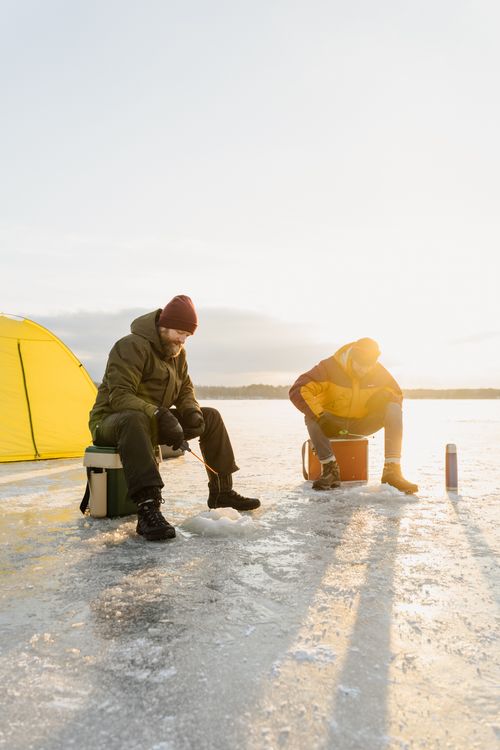 Free Two men ice fishing on a frozen lake at sunrise, surrounded by a winter landscape. Stock Photo