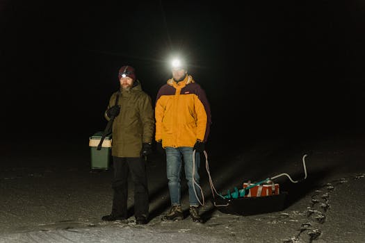 Two men ice fishing at night, equipped with headlamps and gear on a snowy landscape.