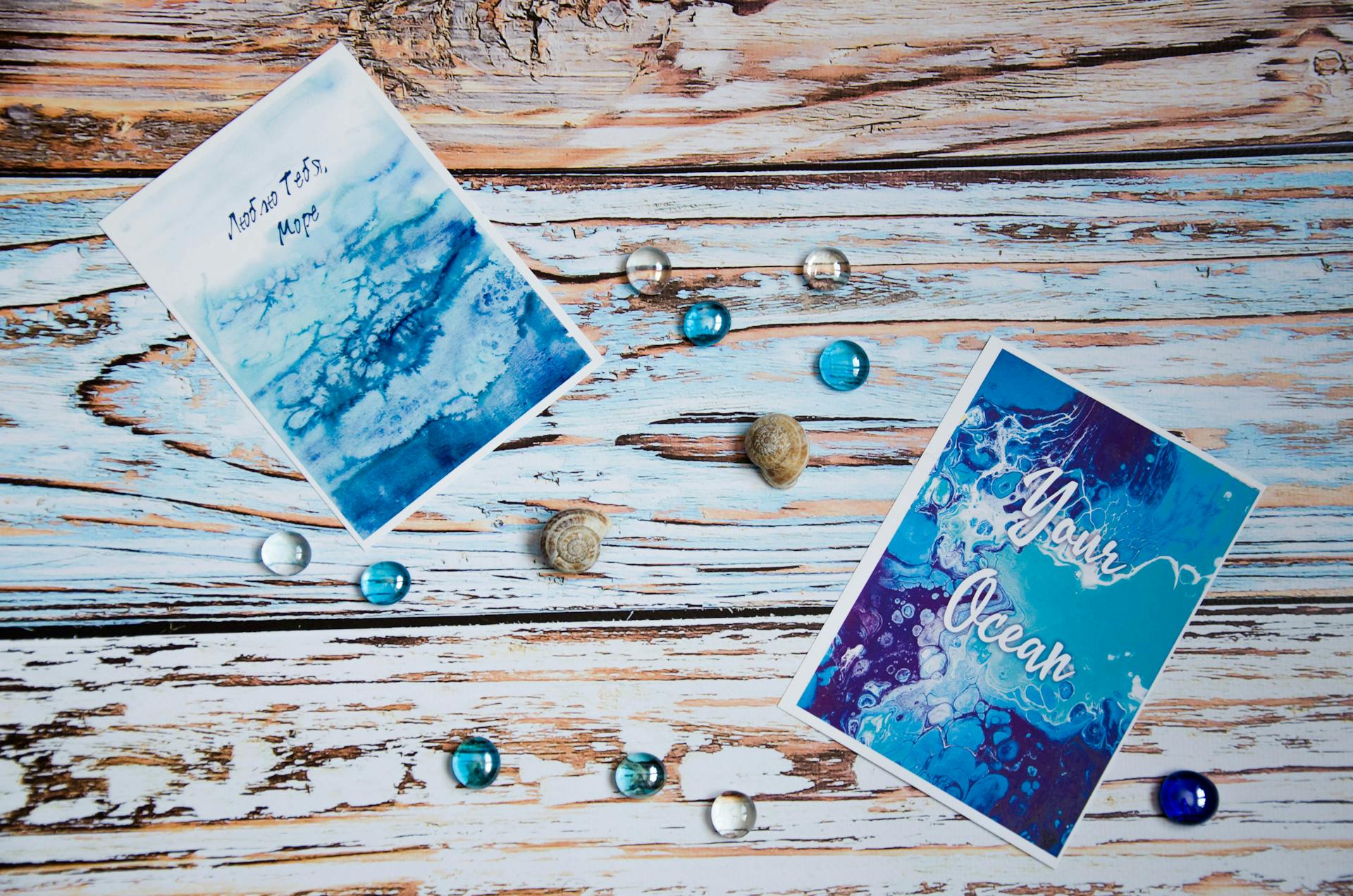 Two artistic ocean-themed postcards surrounded by decorative shells and gems on a rustic wood surface.