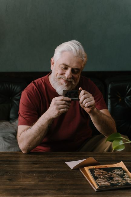 A senior man with a beard smiling while holding a cassette tape, enjoying a moment of nostalgia indoors.