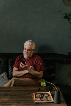 A senior man with white hair using a mobile phone while sitting on a dark couch indoors.