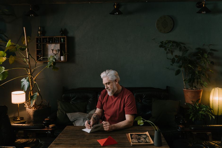 An elderly man writing thoughtfully on a card at a cozy, plant-filled table
