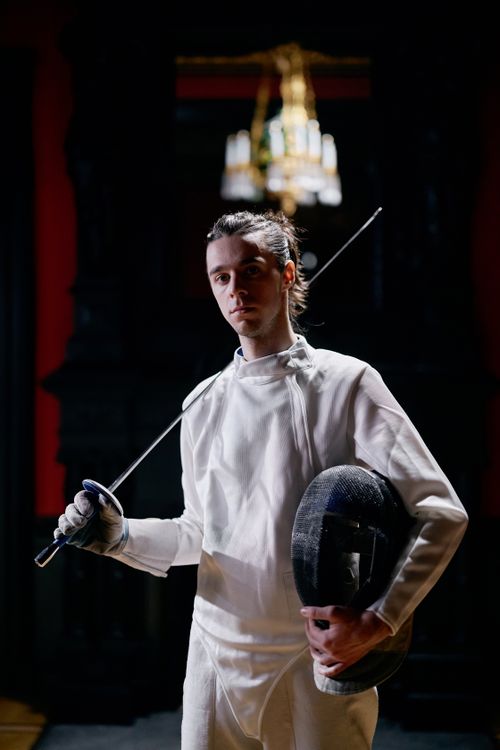Free Adult male fencer standing indoors in full fencing gear, holding a sword. Stock Photo