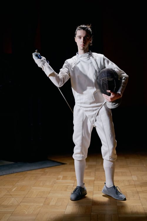 Free Caucasian male fencer in full gear holding a foil and mask, standing indoors. Stock Photo