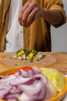A man sprinkles herbs while making fresh spring rolls indoors.