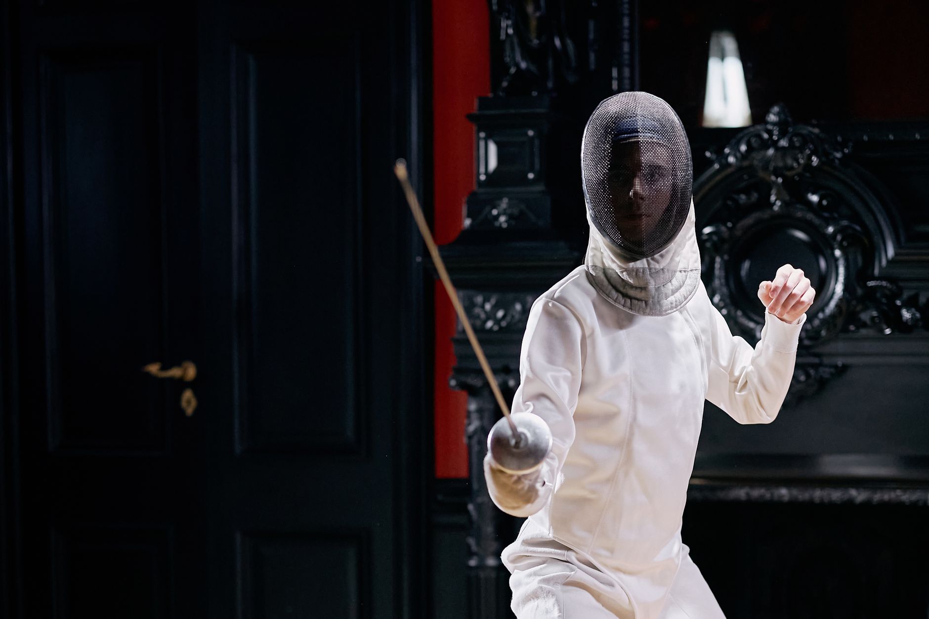 A fencer in a white uniform practicing indoors with focused stance and foil.