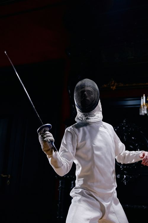 Free A fencer in full uniform striking a pose with a foil indoors. Stock Photo