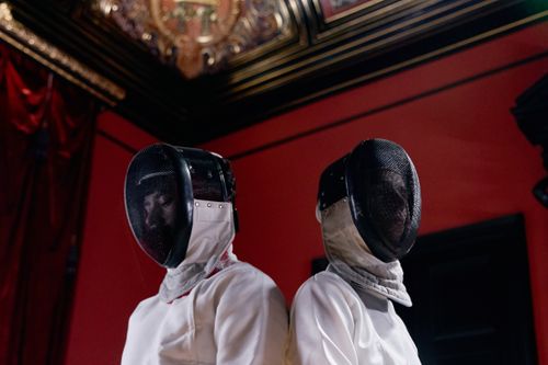 Free Two fencers in gear stand back-to-back in an ornate, dimly lit room. Stock Photo