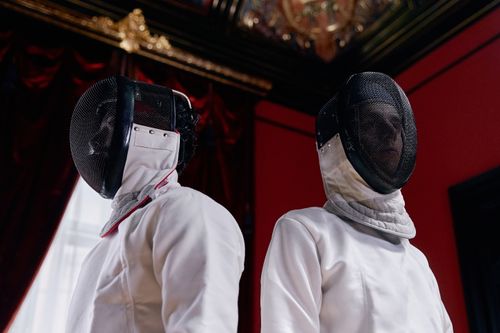 Free Two fencers wearing helmets and white outfits ready for a duel in a classic indoor setting. Stock Photo