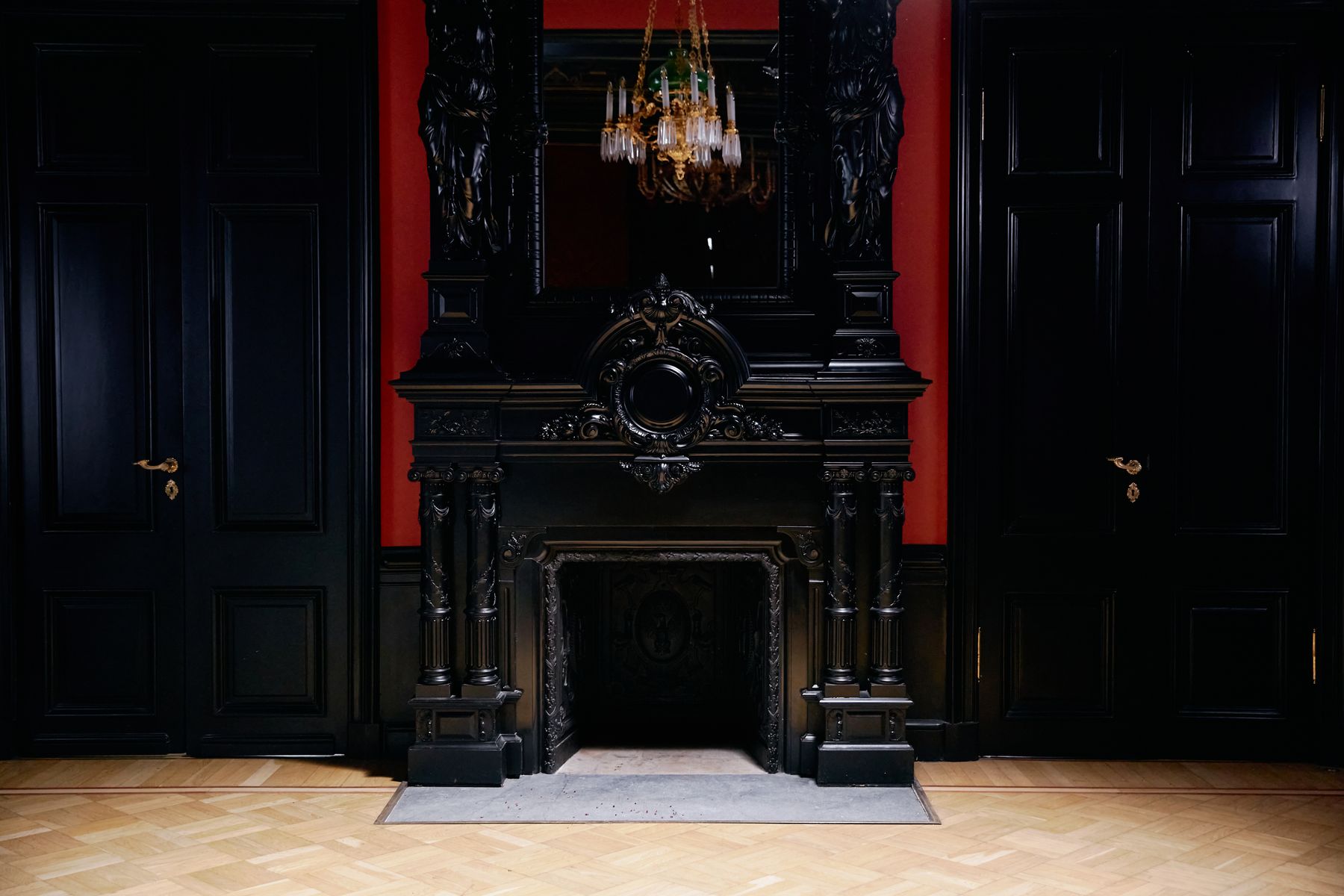 Ornate black fireplace between paneled doors with chandelier and red upholstered chair