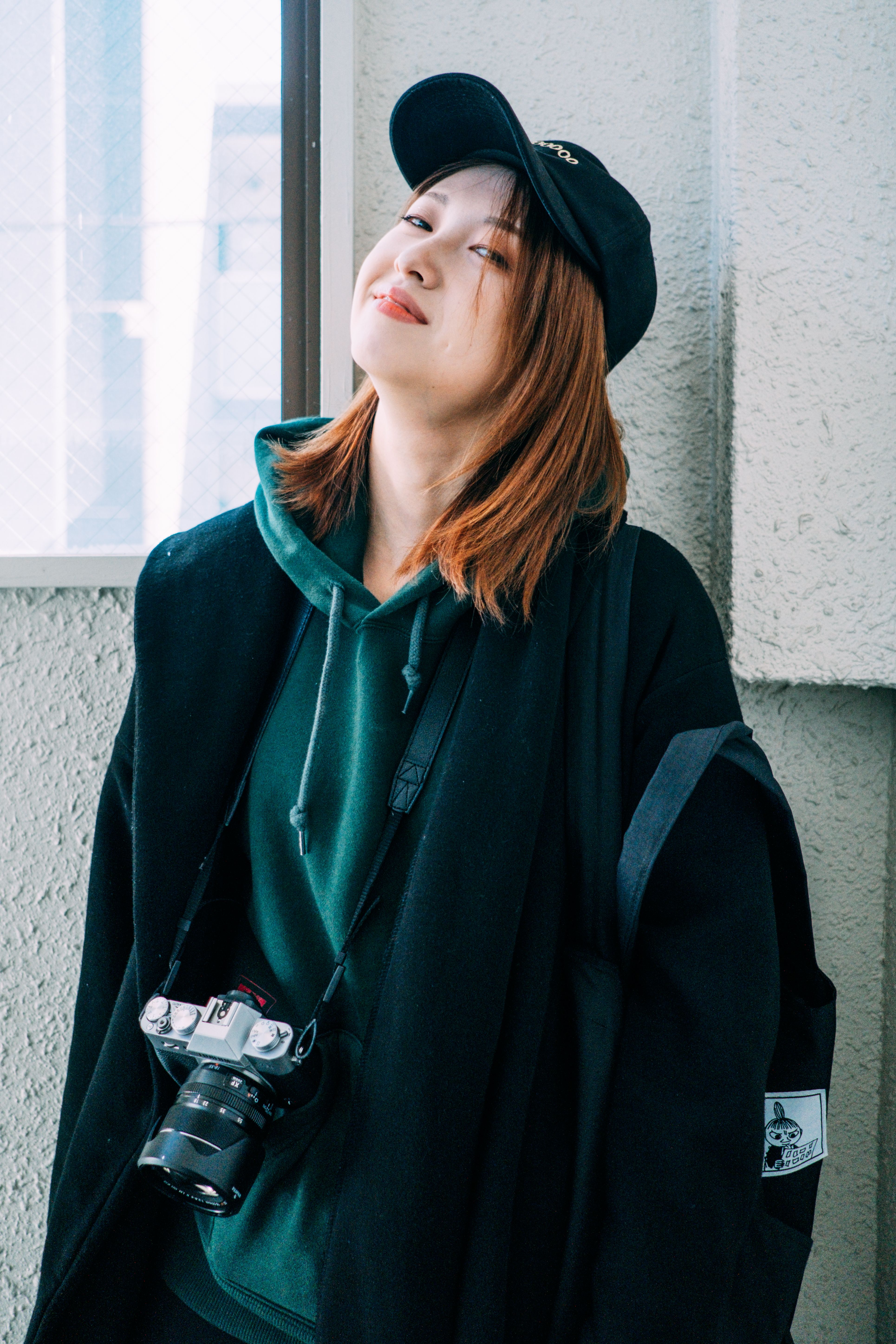 Positive young Asian female millennial in trendy outfit and cap with photo camera on neck smiling and looking at camera while standing near concrete wall in city
