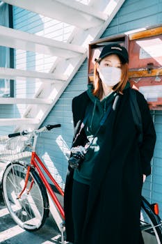 Trendy young Asian female traveler with red hair in stylish outfit and face mask standing near parked bicycle and aged residential house on sunny day during coronavirus