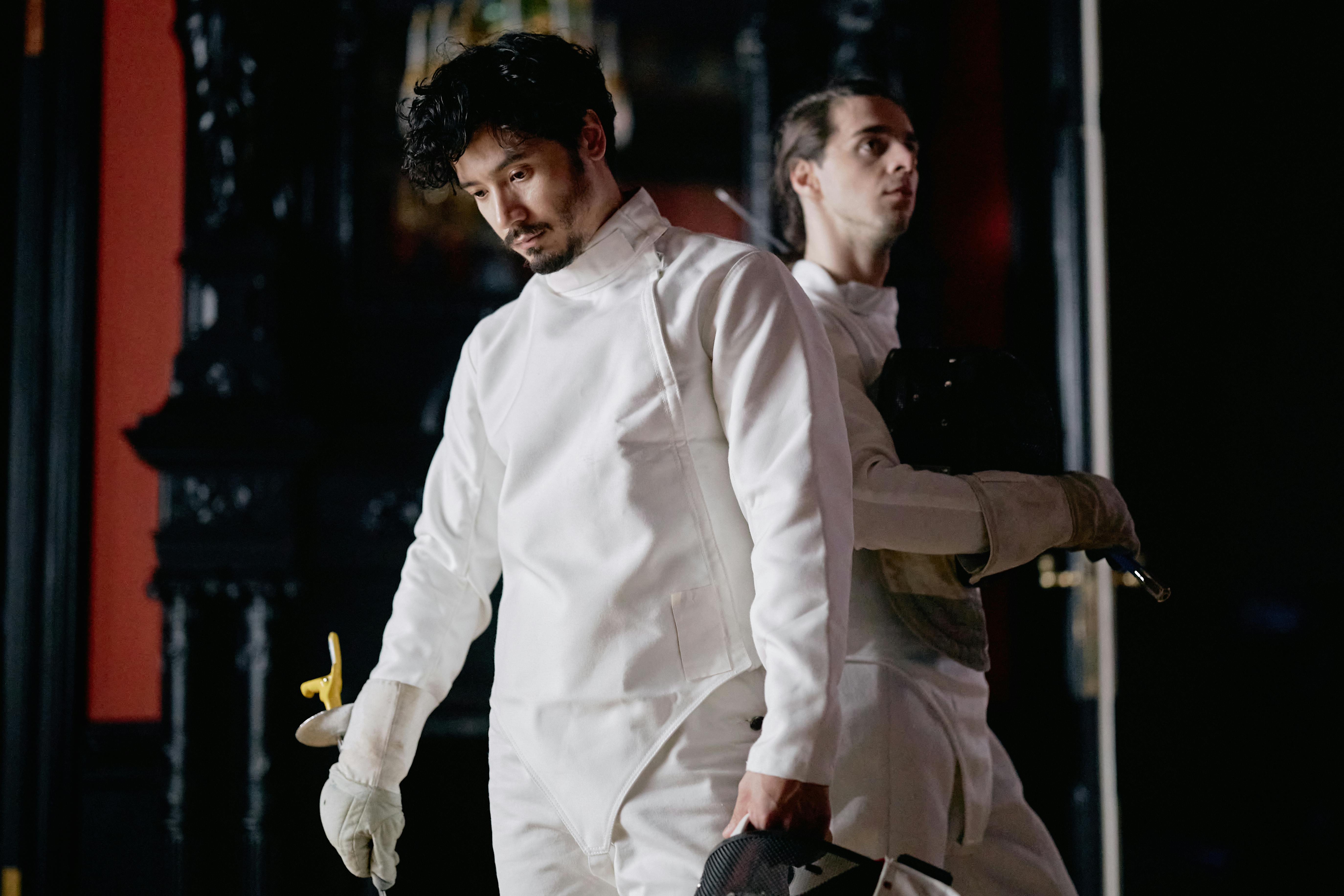 Two male fencers in white gear focus before a match indoors.