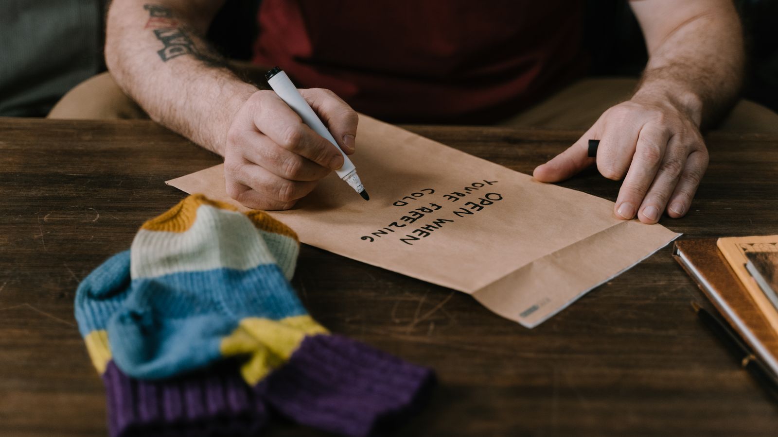 Hands writing an open-when note on a brown envelope beside a colourful knitted garment