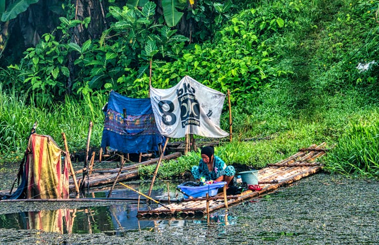 A Woman Washing Clothes In The River Sitting On A Bamboo Raft