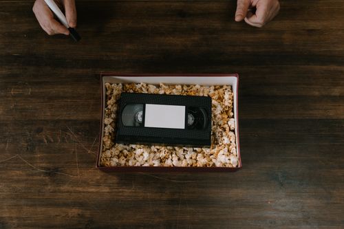 Free A VHS tape surrounded by popcorn in a box, placed on a wooden surface. Hands holding a marker are also visible. Stock Photo