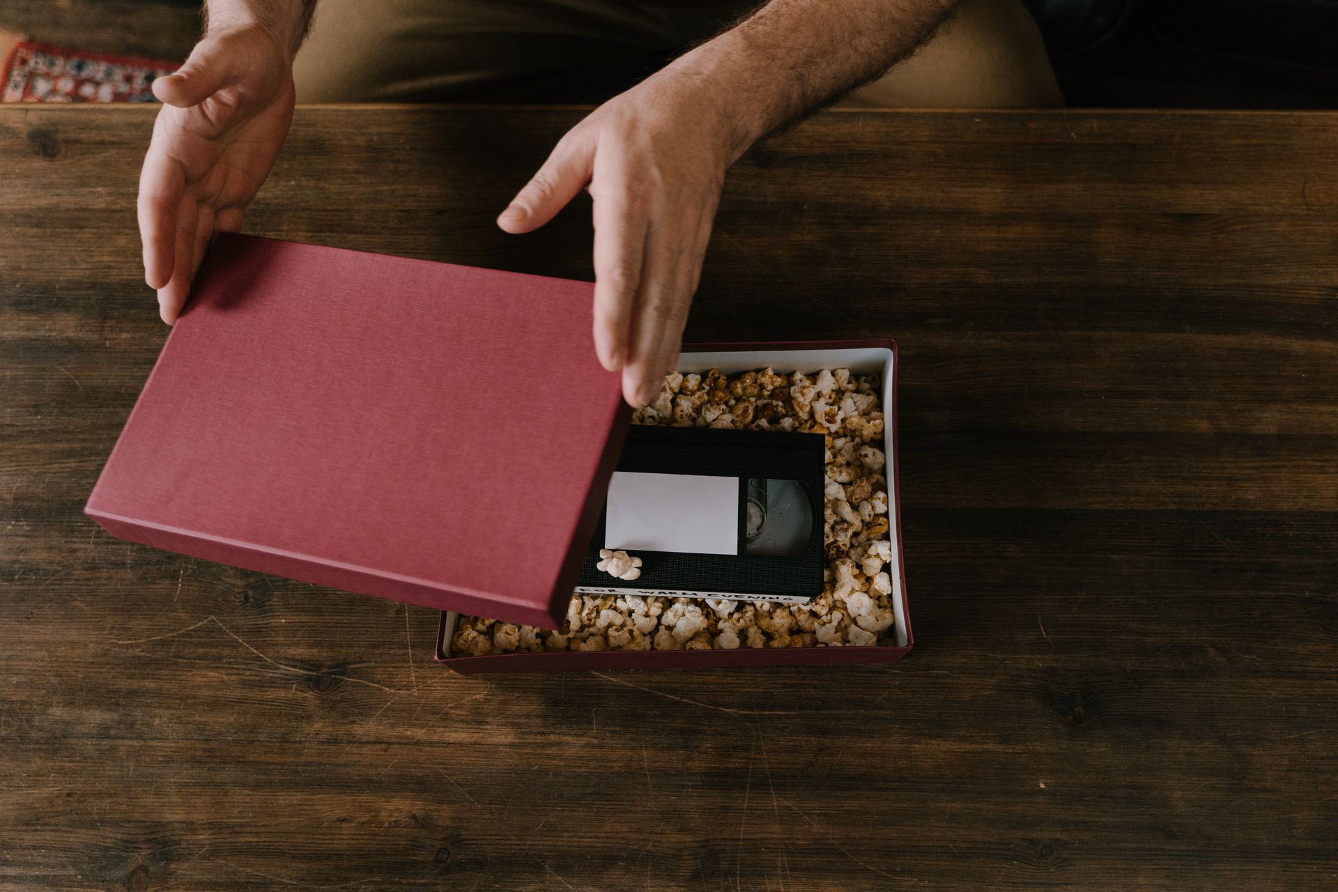 Hands opening a maroon gift box containing a VHS tape cushioned in popcorn.
