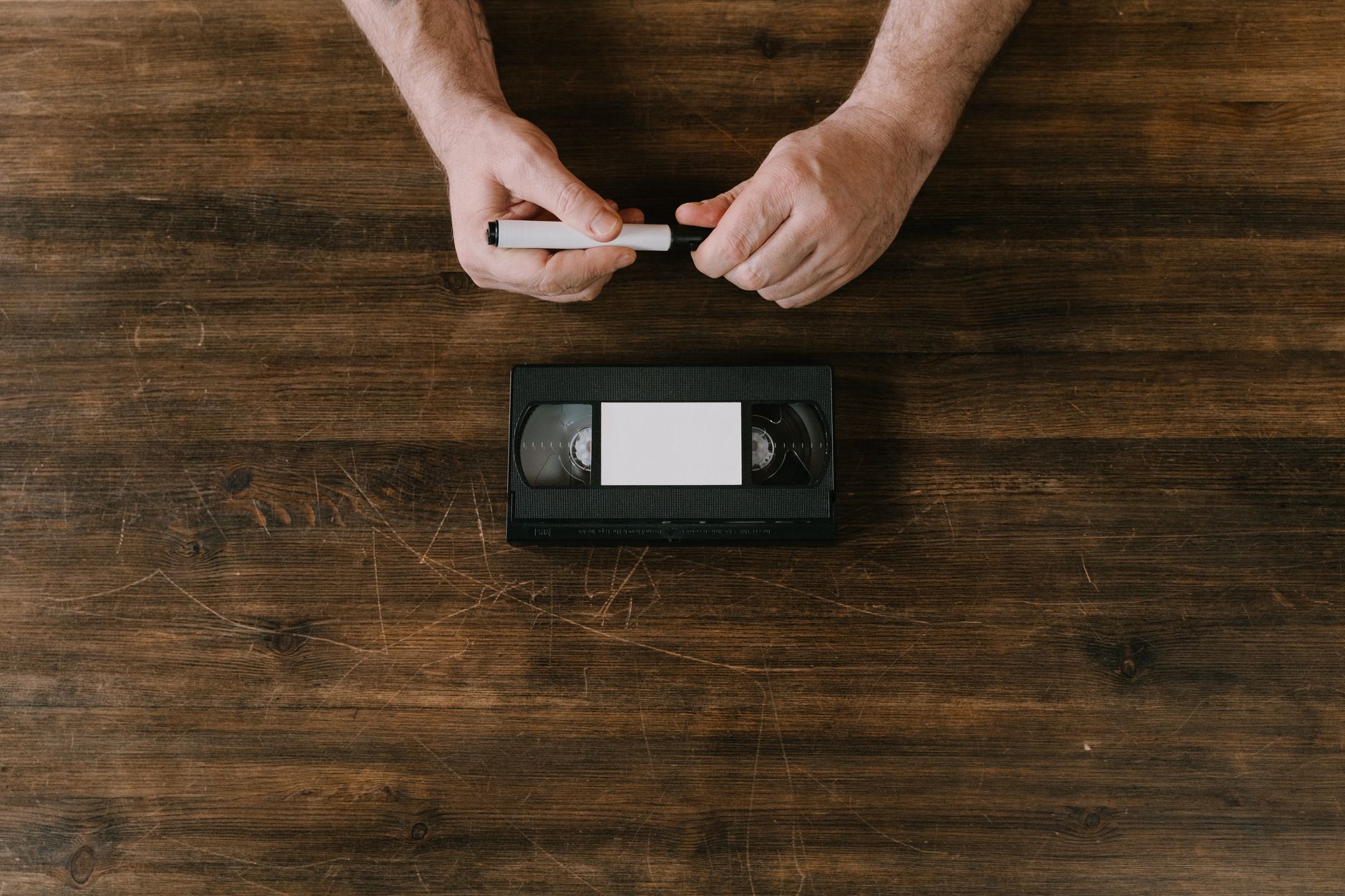 Hands holding marker above VHS tape on wooden table, creating nostalgic vibe.