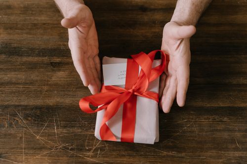Free A beautifully wrapped gift with a red ribbon on a wooden surface, presented by hands. Stock Photo