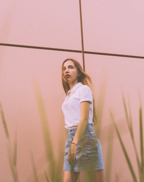 Low angle of self assured young female millennial with long red hair in trendy outfit walking on city street near pink wall