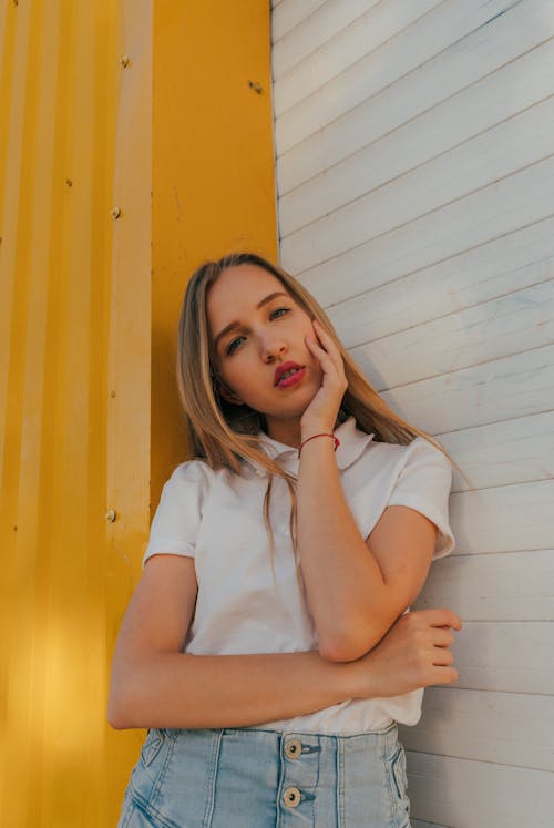 Pensive female teenager with long blond hair in casual outfit touching face and looking at camera with interest while leaning on wall of building on street