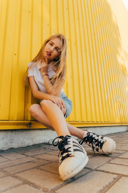 Full body of positive young female millennial with long blond hair in trendy outfit touching chin and looking at camera while sitting on street near yellow metal wall