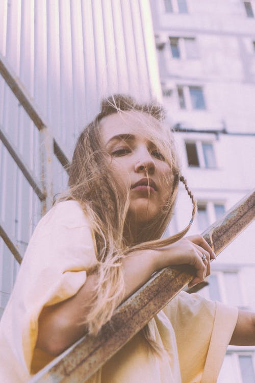 From below of young calm lady with long blond hair with braids leaning on aged metal railing and looking at camera while standing on staircase near multistory building on street