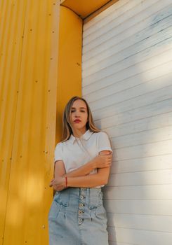Serious young female millennial with long blond hair in stylish outfit looking at camera while standing near steel roller shutter door with crossed arms