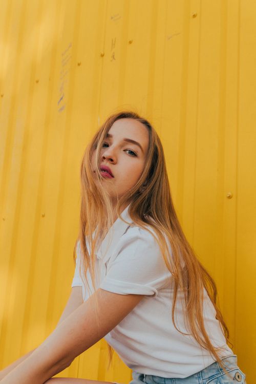 Free Side view of confident young woman with long blond hair in white polo shirt sitting on street near metal yellow wall and looking at camera on sunny day Stock Photo