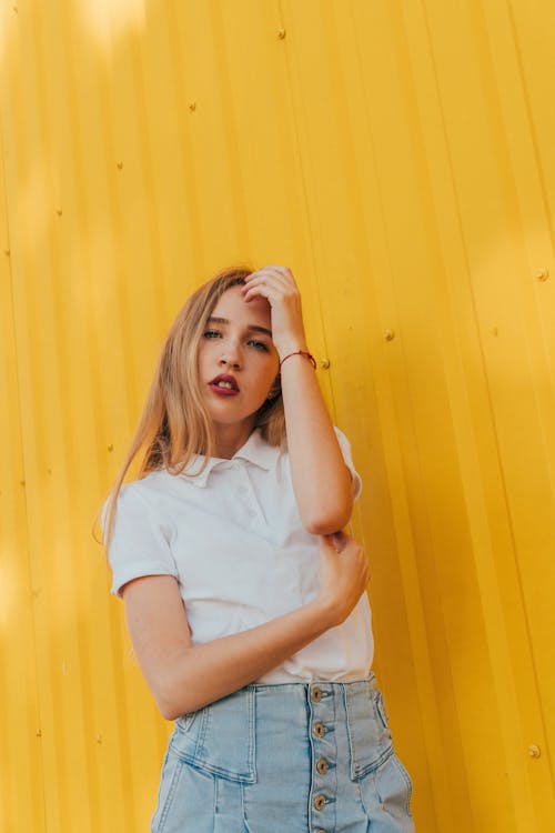 Self esteem young female millennial in stylish casual outfit touching long blond hair and looking at camera while standing near metal yellow wall on street in sunlight