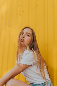 Side view of alluring young lady with long blond hair in white t shirt sitting near metal yellow wall and looking at camera while resting on street