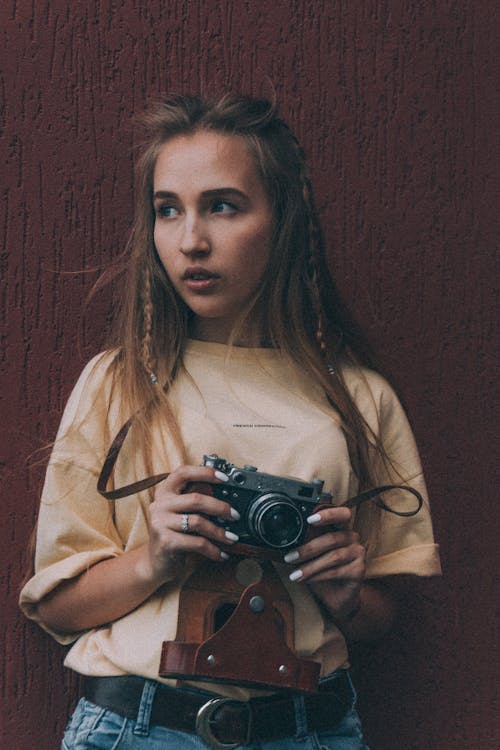 Thoughtful young female millennial with ling bond hair in stylish outfit using vintage photo camera and looking away against brown background