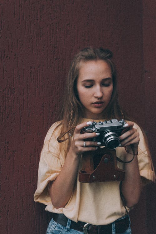 Stylish young female photographer with long hair in casual clothes standing near brown wall and checking pictures on vintage camera