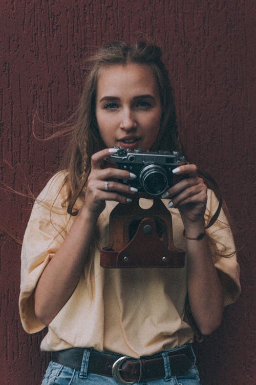 Positive young lady with long hair in stylish outfit smiling while taking pictures on vintage photo camera against brown background