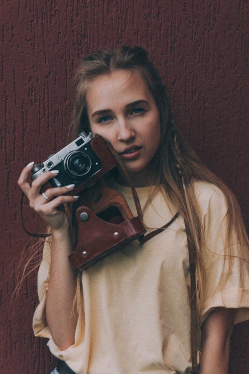 Confident young woman standing near brown wall with vintage photo camera in hand