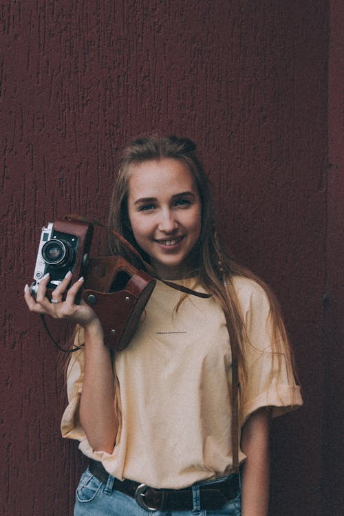 Cheerful young female millennia in stylish clothes smiling while standing near brown wall with vintage photo camera in hand
