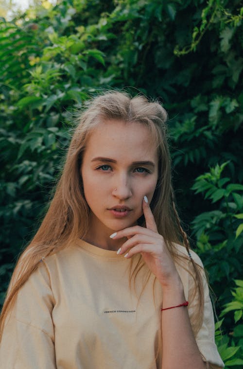 Thoughtful young female model with long blond hair touching cheek and looking at camera while standing against lush green foliage in garden on sunny day